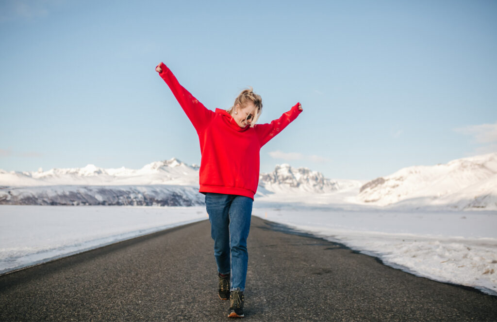 Young, excited woman in red sweatshirt and sunglasses walking down the road with snowy mountains behind her. 